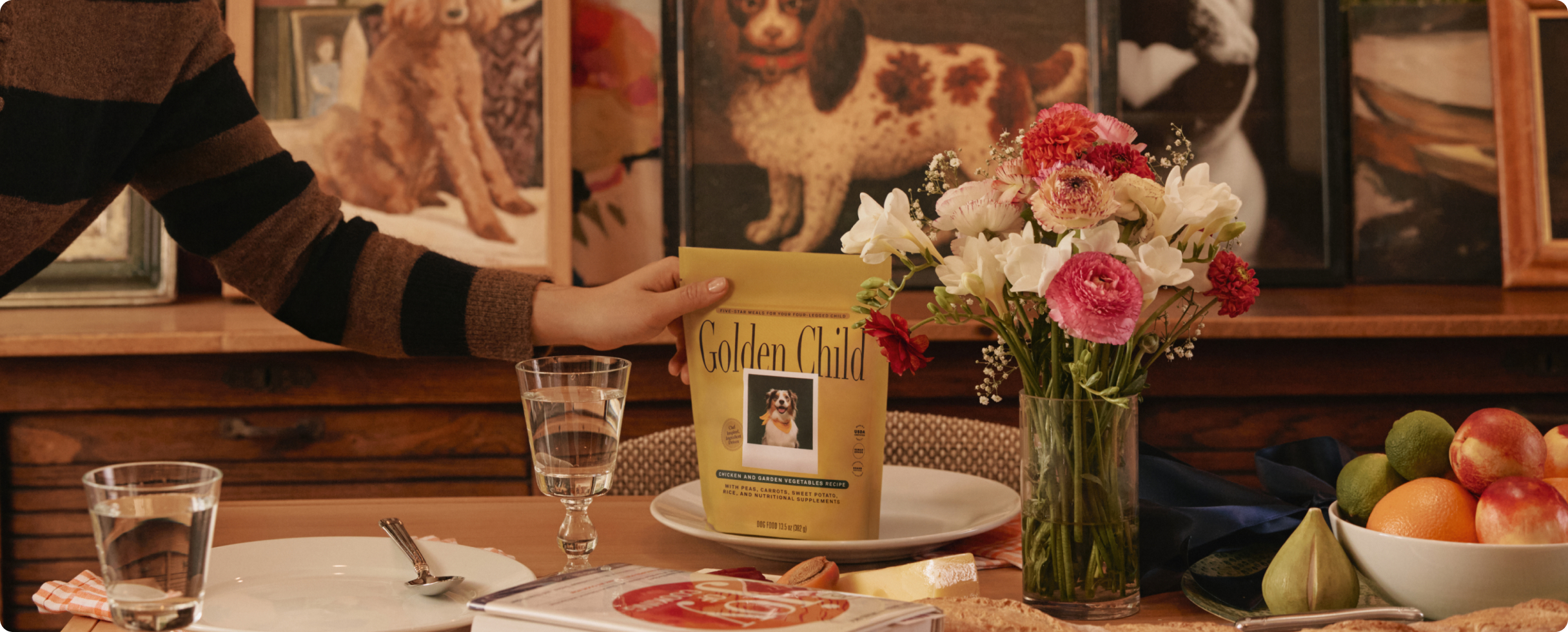 Hand placing a Golden Child fresh dog food pouch on a dinner table set with flowers and fruit